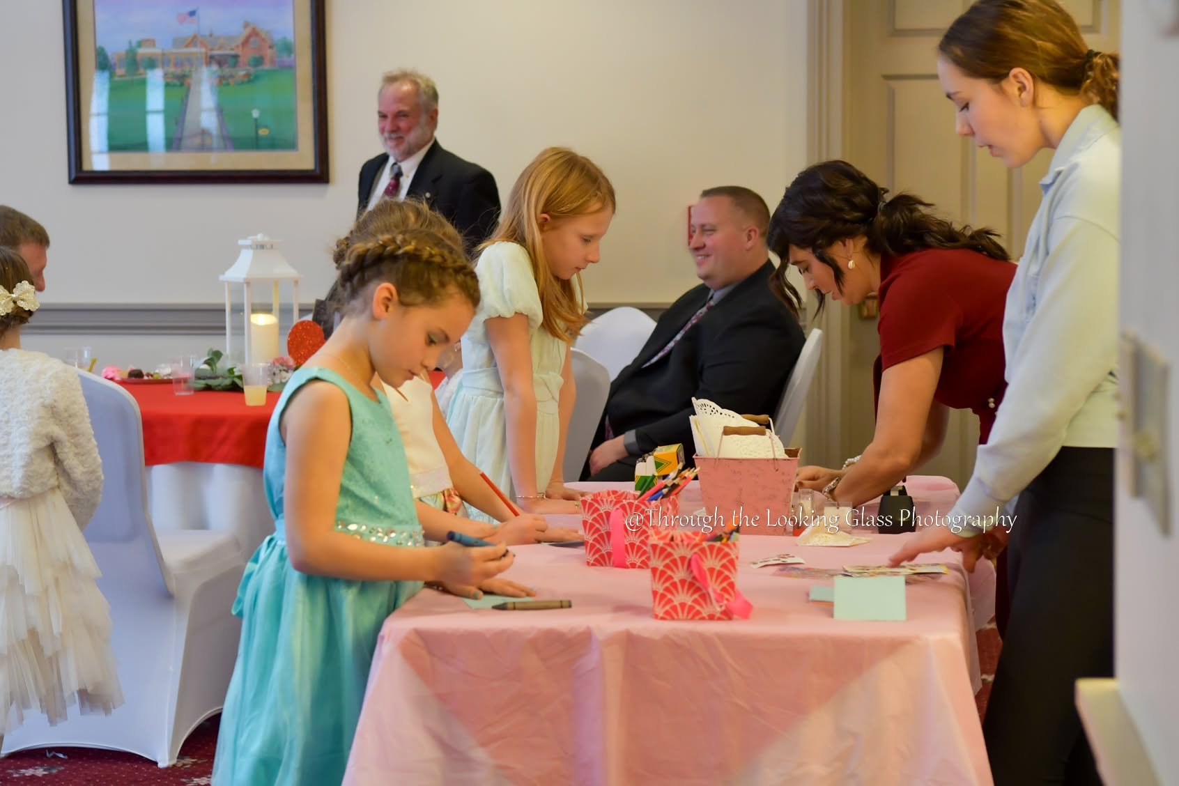 Children and adults at a reception table signing guest book or making cards at a celebration event in a decorated room.