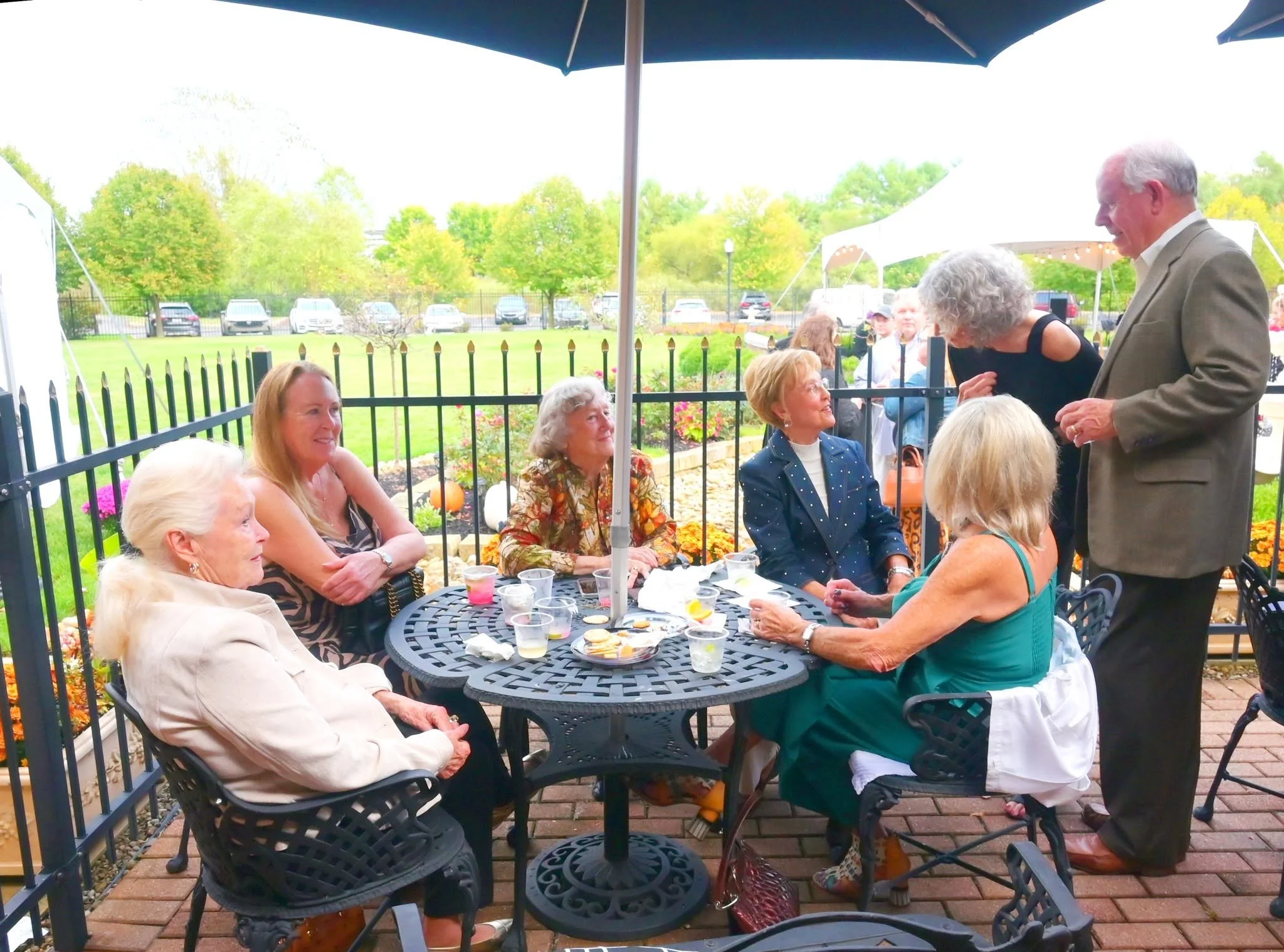 Group of elderly women sitting around a black metal table outside, with one man standing, engaged in conversation, under a large blue umbrella. Green trees and parked cars are in the background.