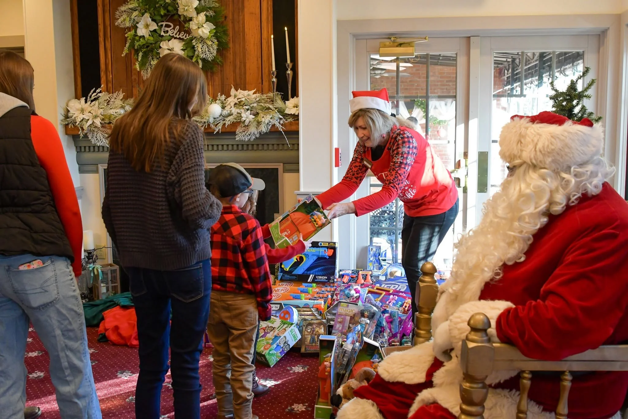 A woman dressed as Santa Claus and children exchanging gifts in a Christmas decorated room with a wreath on the wall and a fireplace.