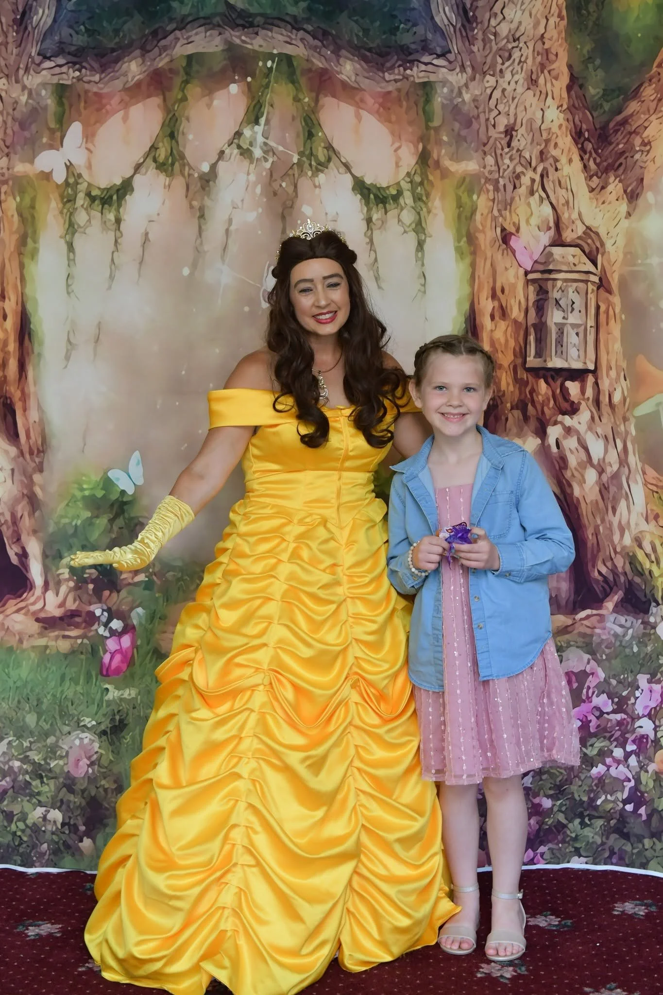 A woman in a yellow dress and a tiara poses next to a girl in a pink dress and denim jacket, standing in front of a fairy-tale backdrop with a tree, flowers, and butterflies.