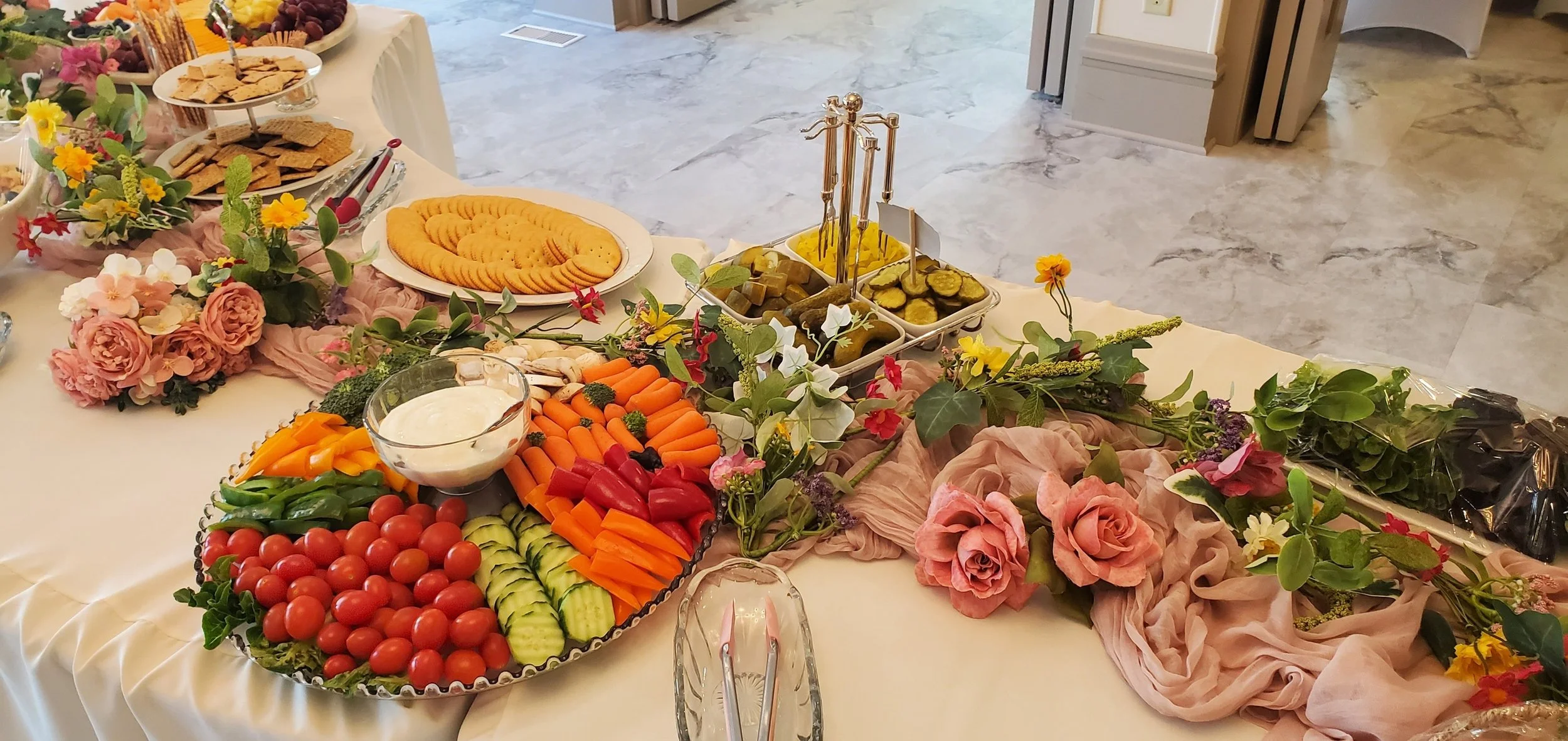 A table at a buffet with various fruits, vegetables, crackers, and desserts, decorated with flowers and pink fabric.