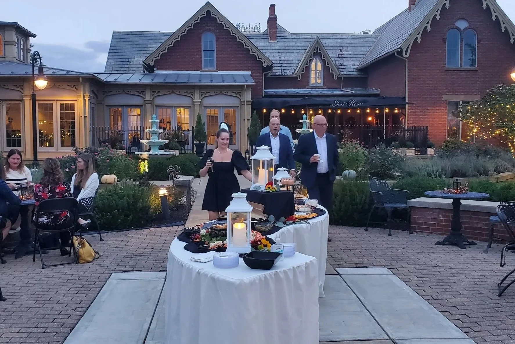 An outdoor evening gathering at a historic house with people mingling, a table with food, lanterns, and string lights creating a festive atmosphere.
