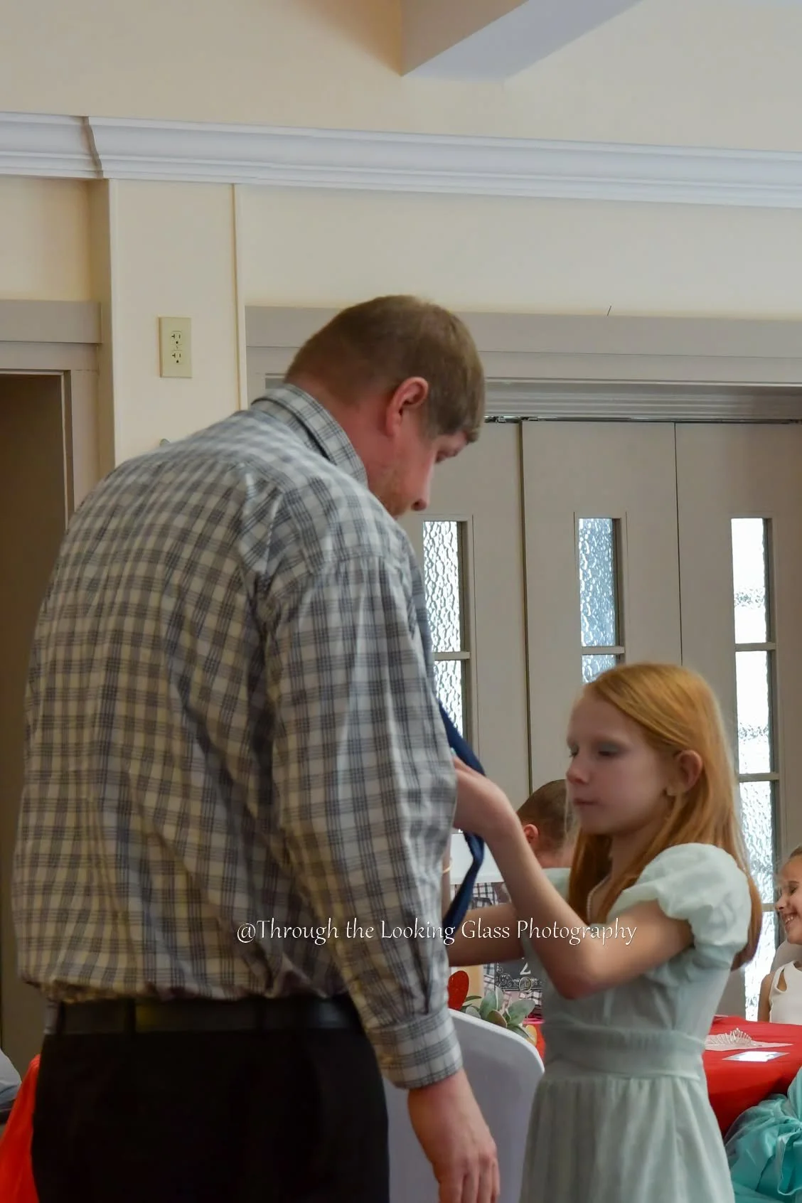 A man in a plaid shirt is helping a young girl in a white dress with a blue ribbon in her hair, into a medal. They are in a room with beige walls, a white ceiling, and a door with glass panels. Several other children are sitting at a table in the background.