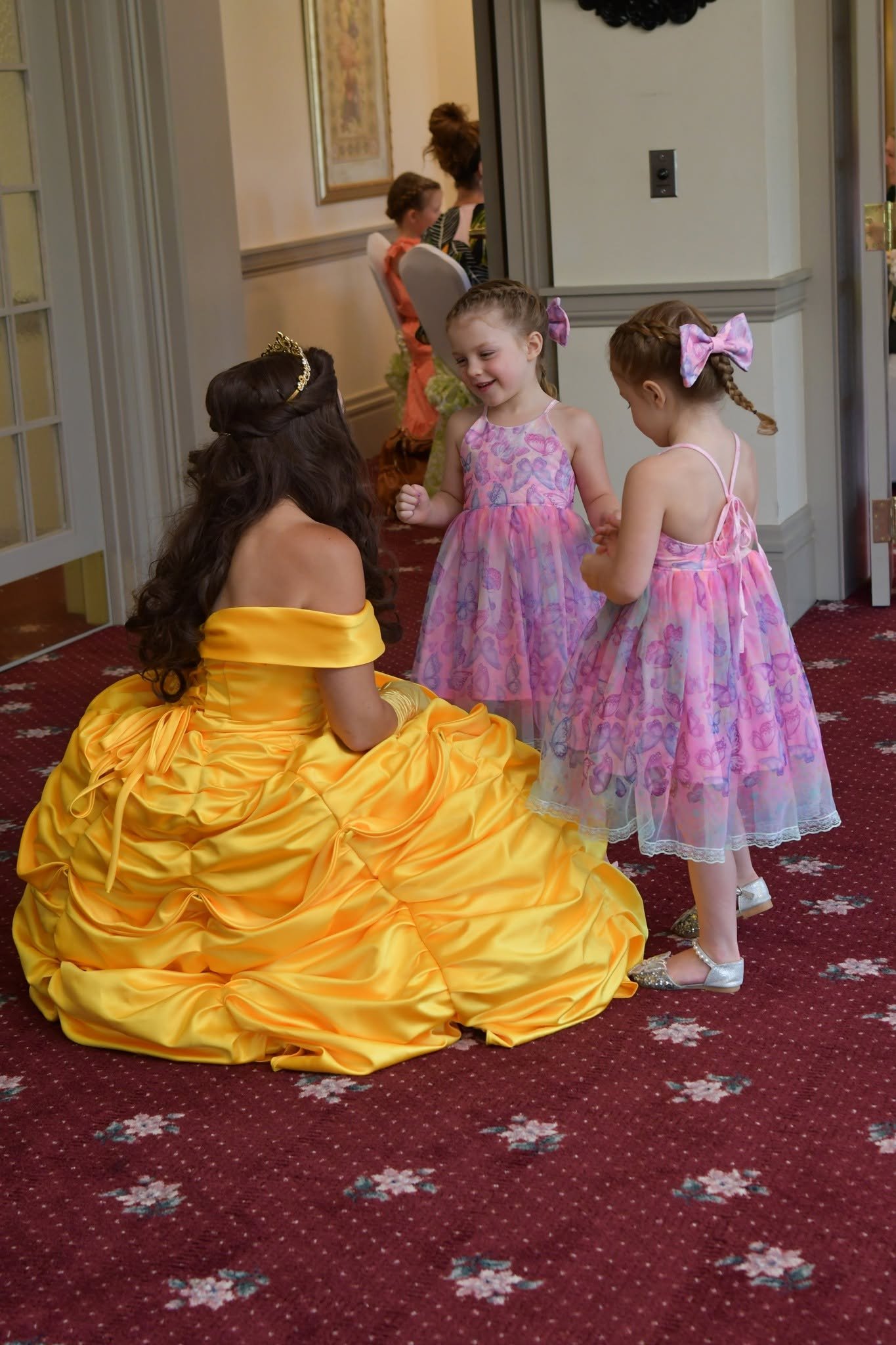 A woman in a yellow gown with a crown on her head is sitting on a red carpet with white and pink flower patterns. She is interacting with two young girls dressed in pink butterfly-themed dresses, one with a pink bow in her hair. The scene appears to be at an indoor event or celebration.