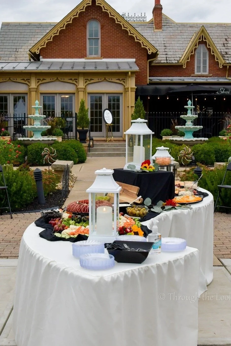 Outdoor event setup with tables of food, lanterns, and decorative elements in front of a historic brick building with ornate trim and fountains.