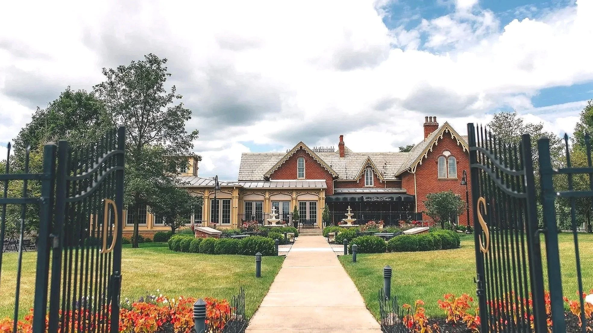 A large house with a red brick exterior and multiple gabled roofs seen through an open black iron gate. The front yard features a stone pathway, well-maintained lawn, flower beds, a fountain, and trees under a cloudy sky.