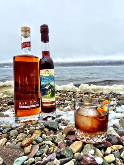 Two bottles of liquor and a cocktail glass with ice and orange peel on a rocky beach.