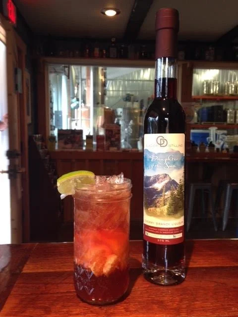 A bottle of Dented Brick Distillery's drink and a glass with a dark mixed beverage garnished with a lime slice, placed on a wooden bar counter.