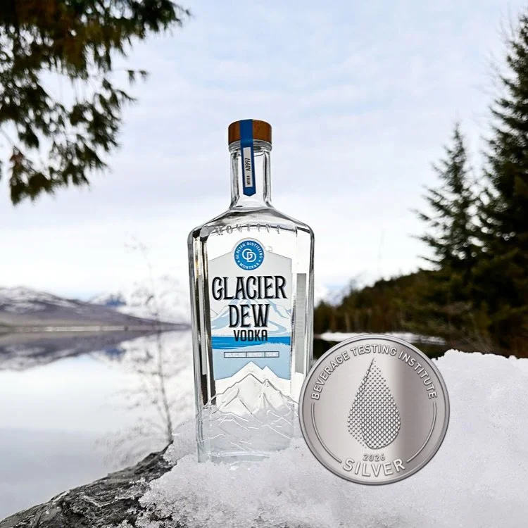 Bottle of Glacier Dew Vodka sitting on a snow covered rock overlooking Lake McDonald in Glacier National Park and greenery in the background. A silver medal is overlaying the photo.