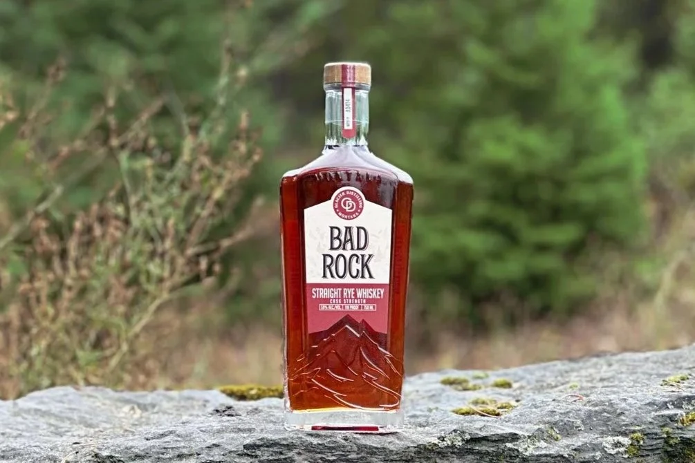 Bottle of Bad Rock sitting on a rock with greenery and fall foliage in the background.