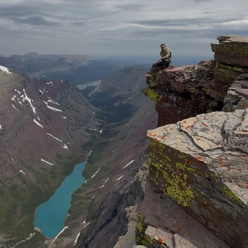 A person sitting on a rocky ledge overlooking a deep valley with a winding river and distant mountains under a cloudy sky.