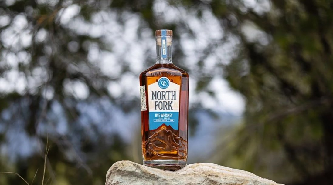 A bottle of North Fork Blue Single Barrel sitting on a rock with greenery in the background.
