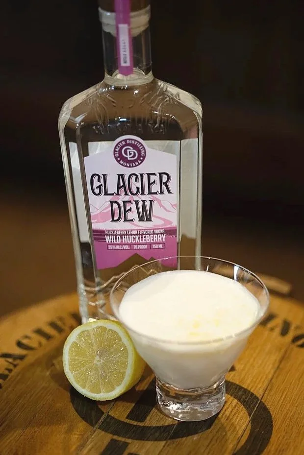 A bottle of Glacier Dew Wild Huckleberry flavored vodka, a glass with a creamy white cocktail, and half a lemon on a wooden surface.