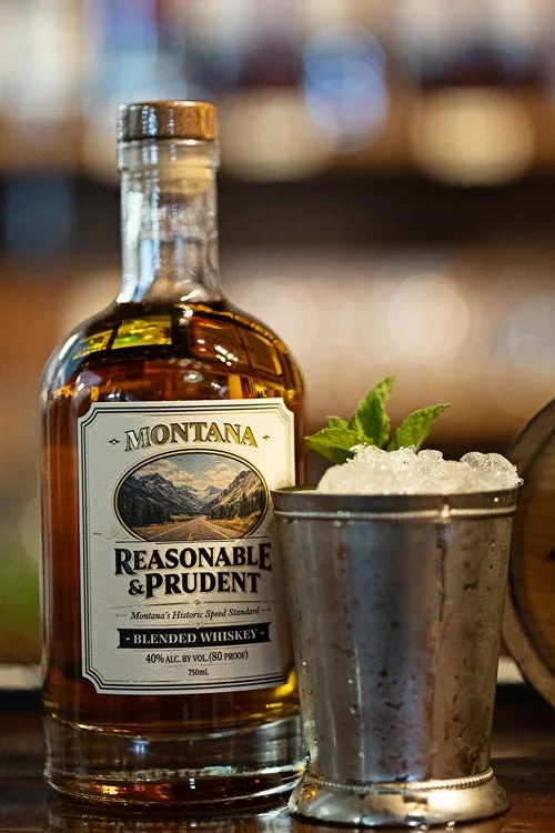 Bottle of Reasonable & Prudent next to a Mint Julep cocktail sitting next to a wooden bar with a part of a small barrel next to it with blurred background.