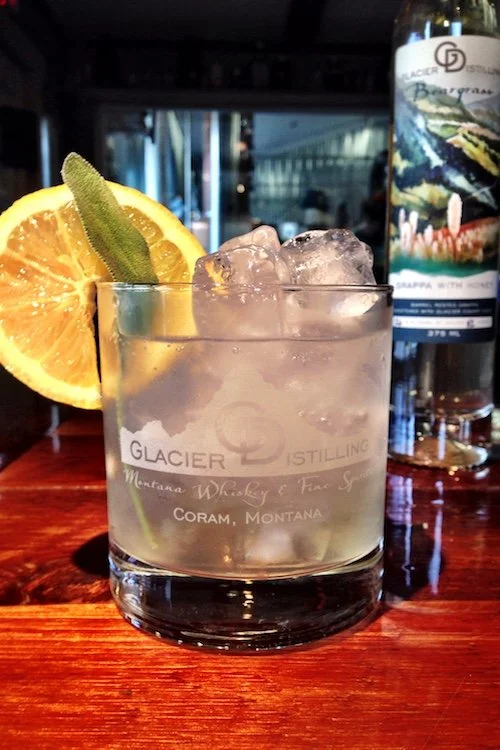 Glass of iced cocktail with lemon slice and sage, branded with Glacier Distilling, alongside a bottle of spirits on a wooden bar.