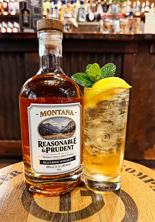 A bottle of Reasonable and Prudent Blended whiskey sitting on a stool with a Montana highball cocktail drink next to it with the Glacier Distilling tasting room bar in the background.