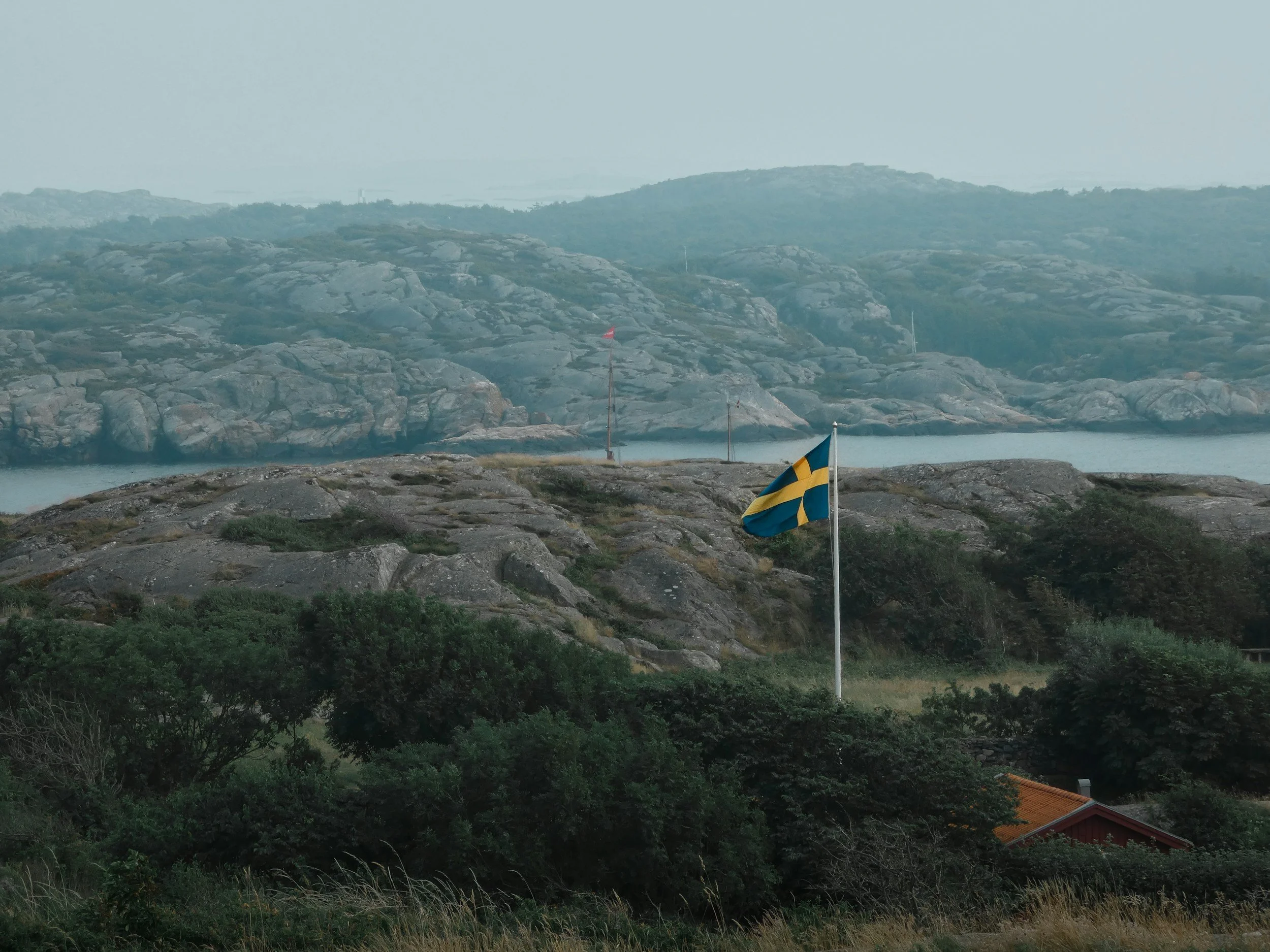 En svensk flagga vajar på en plats med kullar, sten, gröna buskar och ett vatten. I bakgrunden finns fler landformer och vatten.