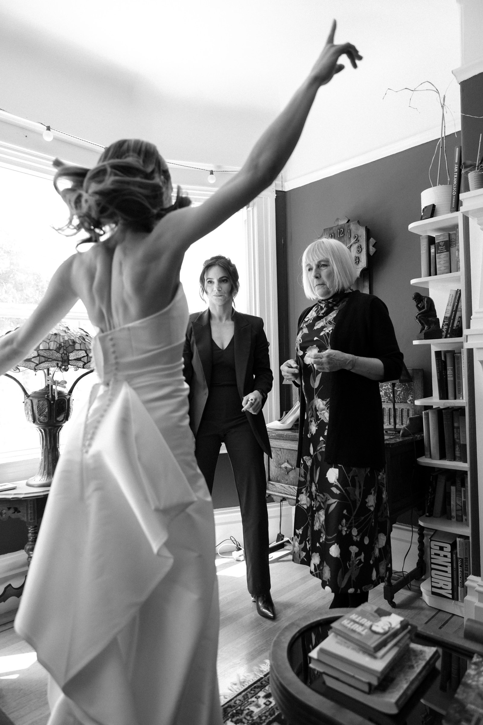 A bride, photographed from behind, jumping up and down right after putting her wedding dress on. Her mom and best friend are watching.