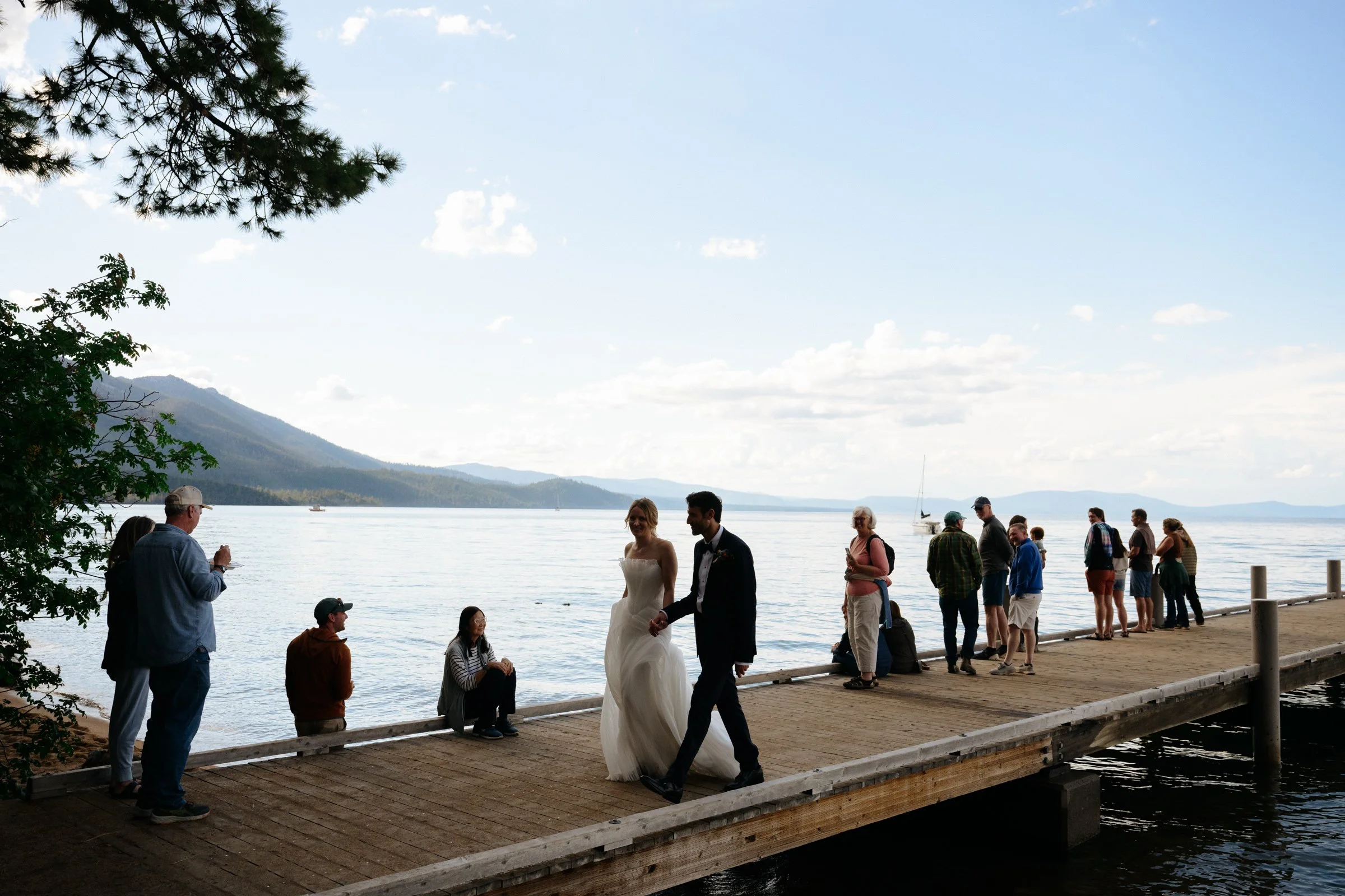 A bride and groom holding hands, walking down a pier on Lake Tahoe, with tourists in casual clothes all around them.
