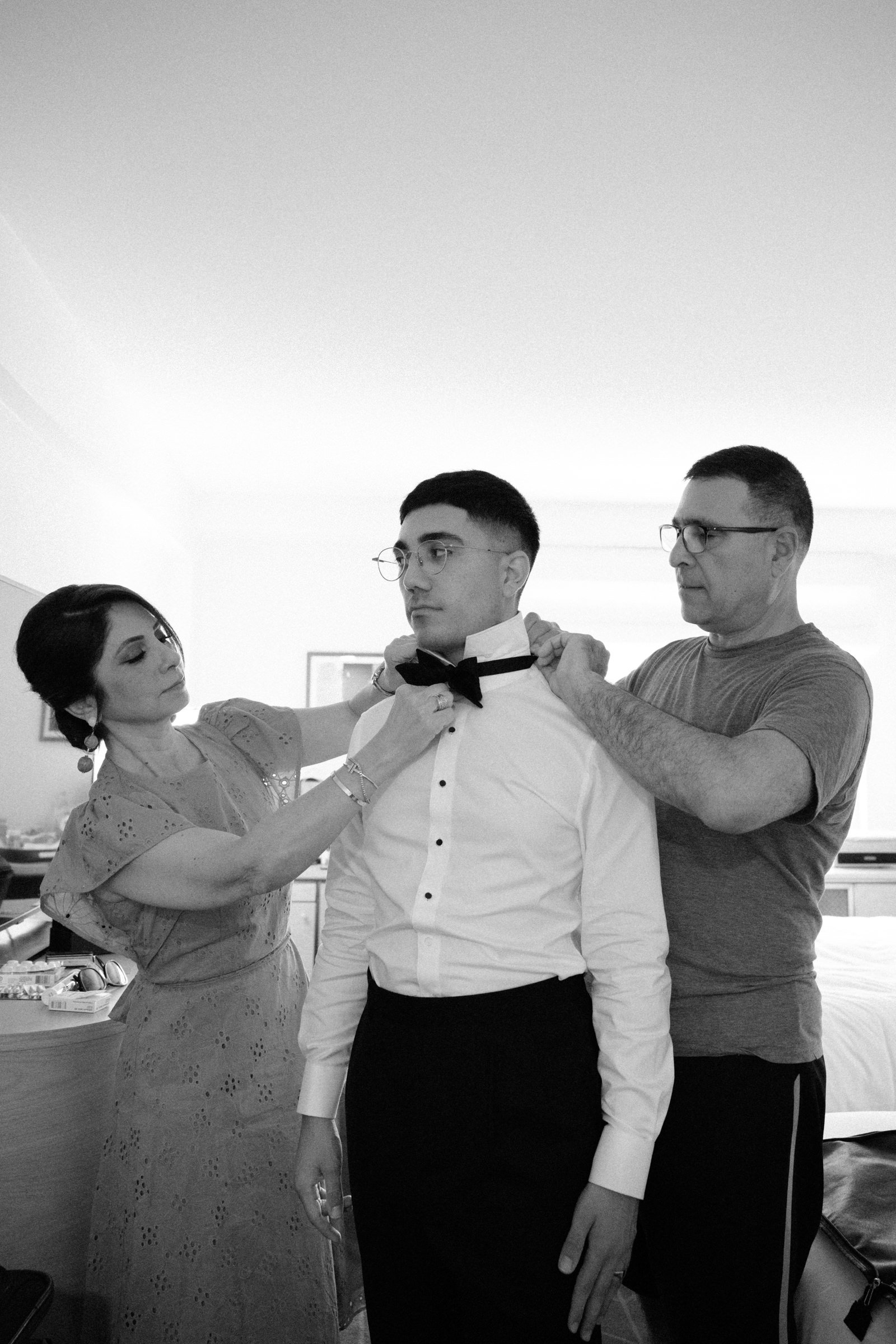 Mother and father of a groom helping him tie his bowtie, before getting married.