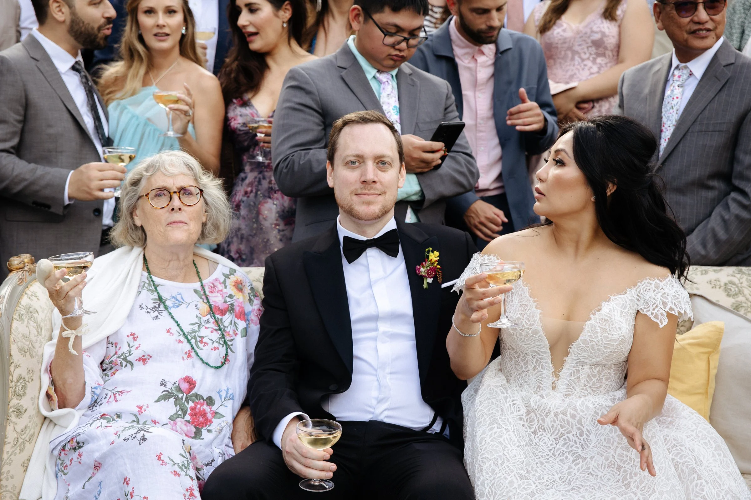 A groom and mother of the groom looking straight at the camera, the bride and others all around them.