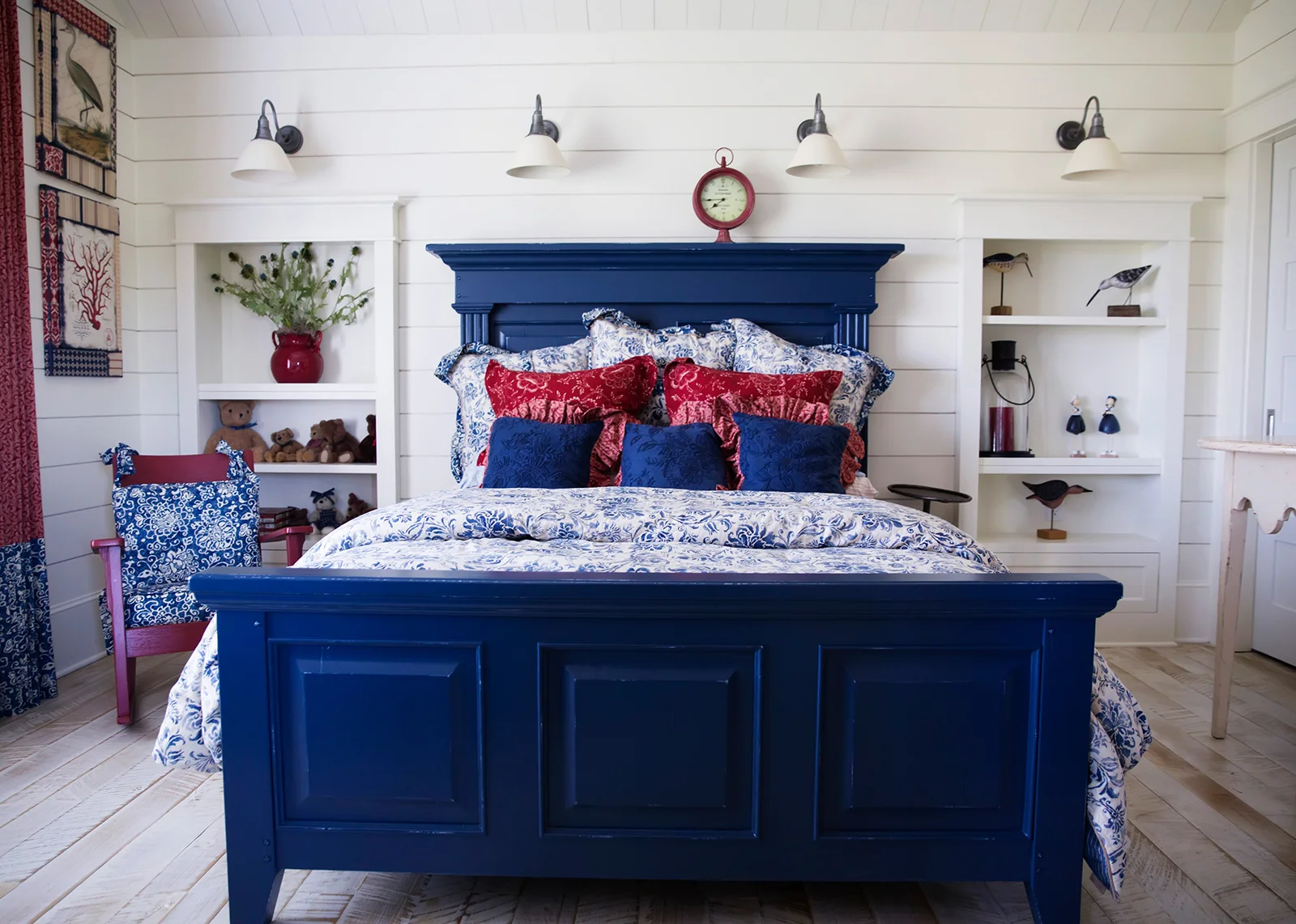 Red and blue bedroom with a quaint, farmhouse look Resources: Bed: Thomasville Furniture Sconces: Restoration Hardware Rocking Chair: Antique Family Heirloom Bedding: Ralph Lauren Home Collection Design Thought: In decorating bedrooms start with a bed skirt, then layer on sheets and a blanket, tucked in together. Mix and match various patterns and colors. Lay a folded duvet across the end of the bed. At the head of the bed pile on the Euro shams and throw pillows in front of the sleeping pillows. 