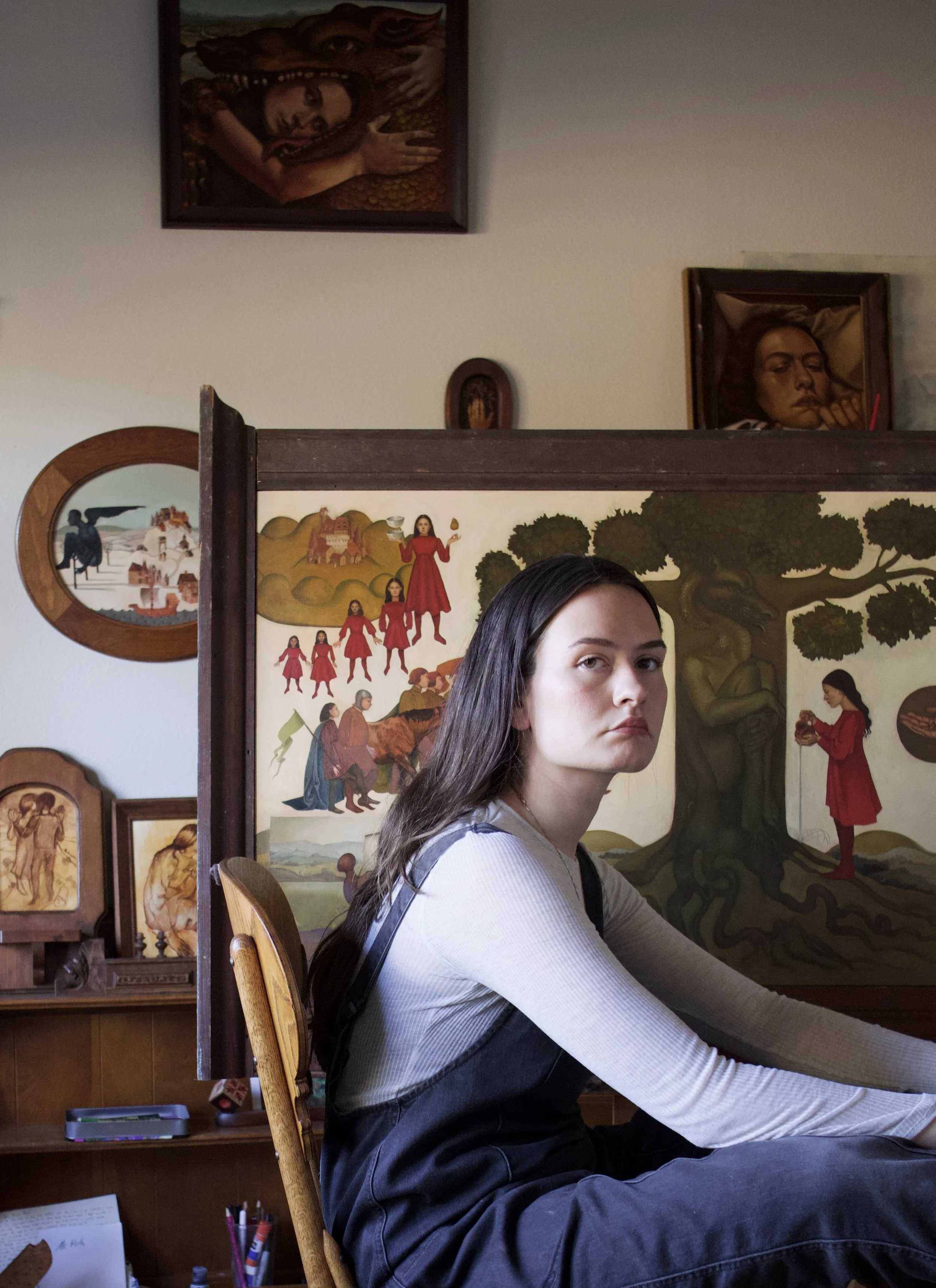 A brunette woman sitting in front of a large painting in a studio. She is wearing black overalls and a grey shirt. The paintings behind her contain portraits of women, dragons, and other creatures.
