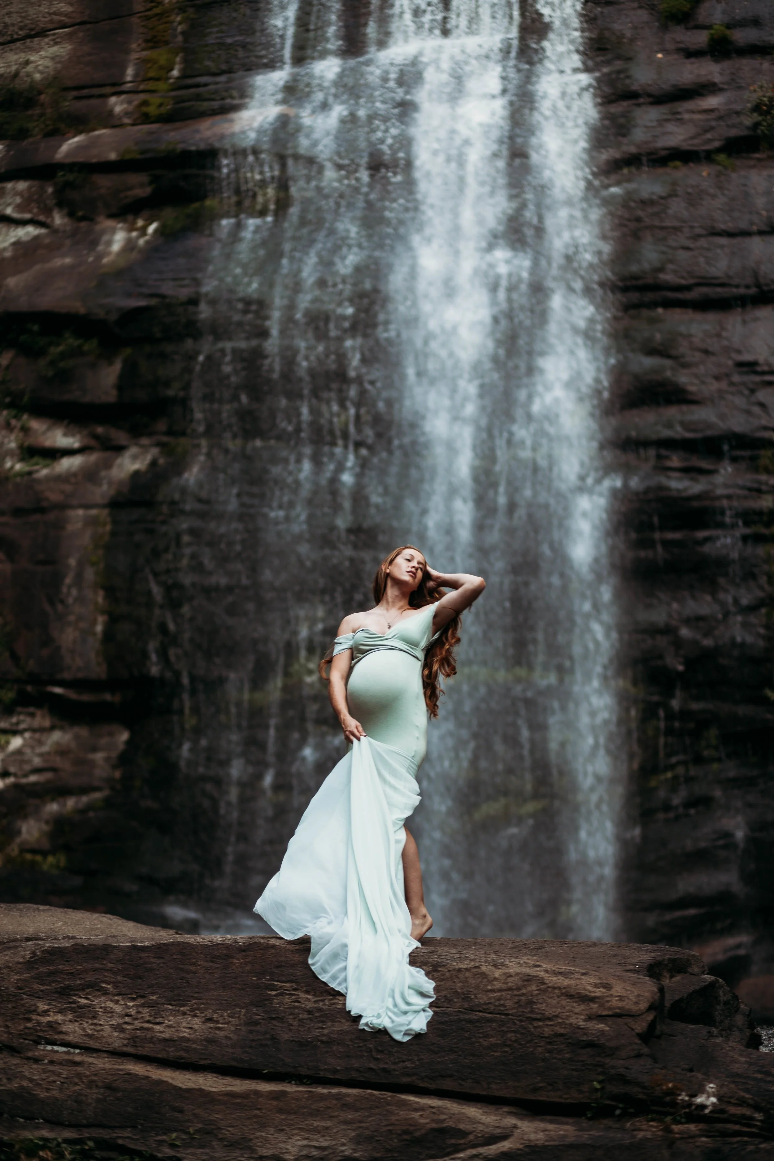 A pregnant woman in a flowing light green dress stands on a rock in front of a waterfall for a maternity session in the Blue Ridge Mountains, with one hand behind her head and the other holding her dress.