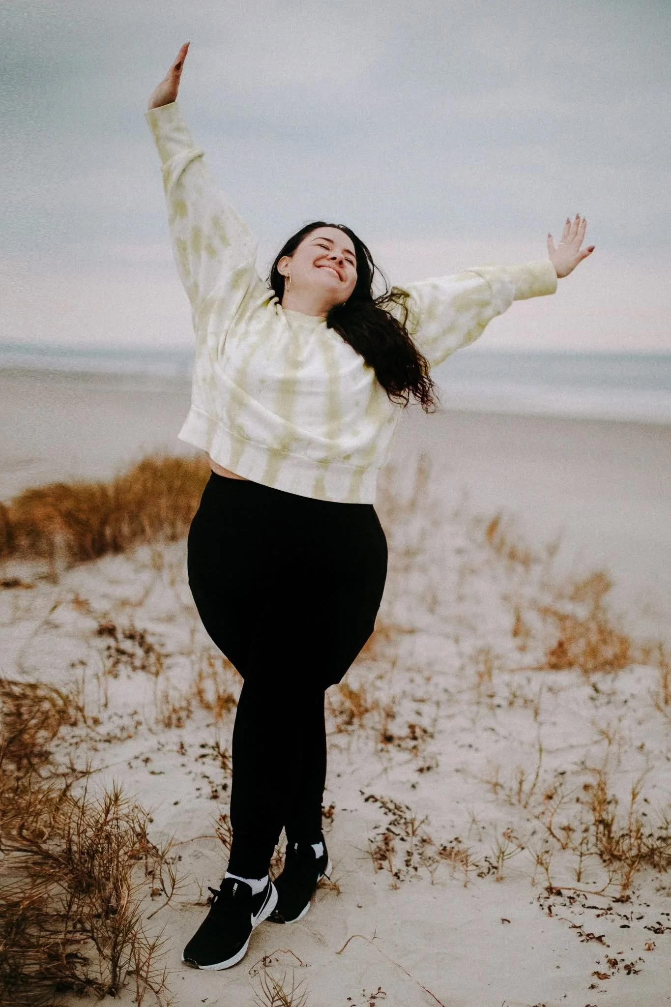 An excited documentary & wedding photographer standing on a beach in Sullivans Island, South Carolina with arms outstretched, smiling, wearing a yellow tie-dye hoodie, black pants, with beach grass and dunes in the background under an overcast sky.