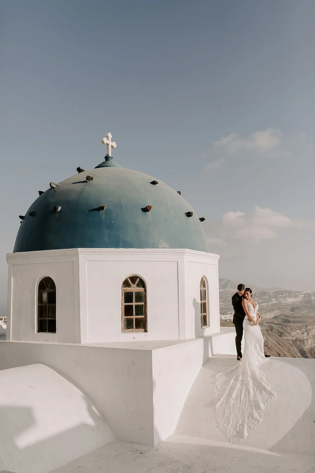 A bride and groom in wedding attire standing outside a white church with a blue dome topped with a cross, in a scenic location with mountains in the background.