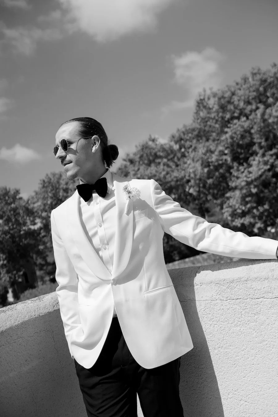 Man in a white tuxedo jacket, black bow tie, sunglasses, with hair tied back, standing outdoors against a wall with trees and a partly cloudy sky in the background.