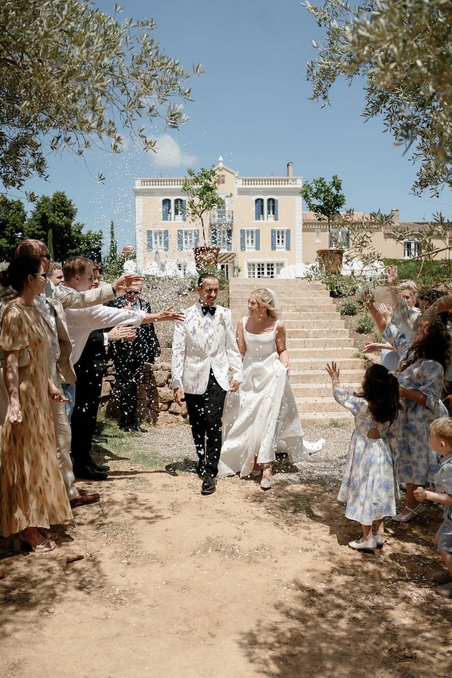 A newlywed couple walks down an outdoor aisle, celebrating their wedding with friends and family throwing rice or confetti. The bride is in a white dress and the groom in a white tuxedo. Guests cheer and throw celebratory items on a sunny day in front of a large, elegant house with stairs and trees.