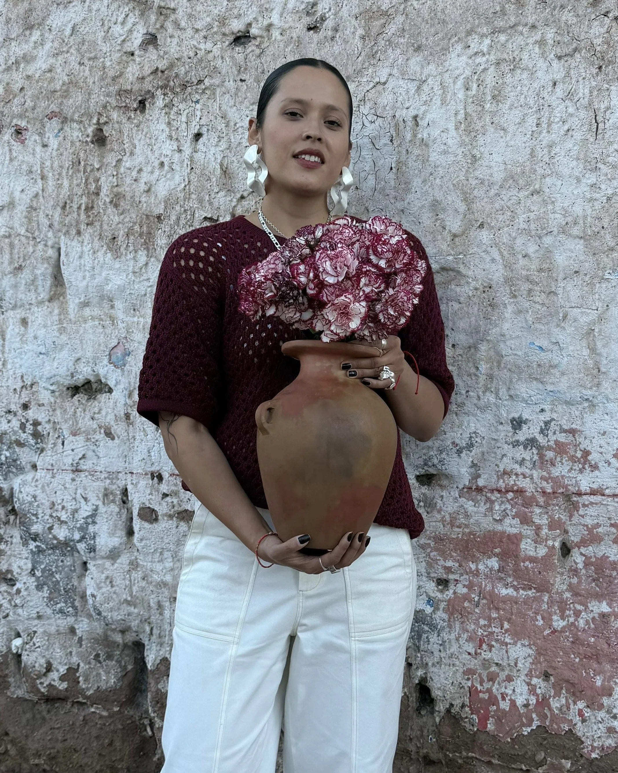 A woman standing in front of a weathered brick wall, holding a large clay vase filled with pink and white flowers, wearing a burgundy open-knit sweater and white pants, with large white earrings and silver jewelry.