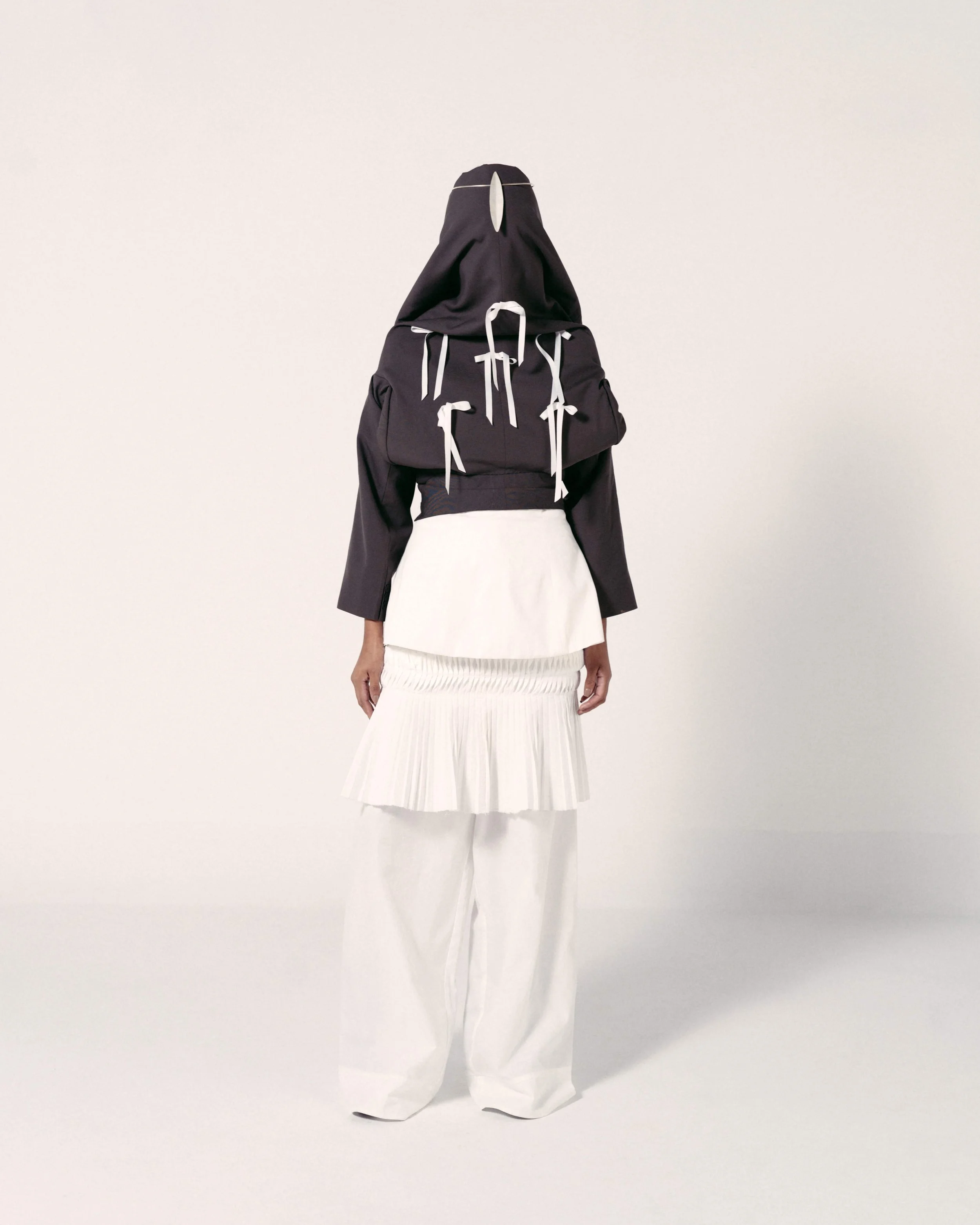 A person wearing detached black and white clothing with a black hooded top with white ribbons, a white skirt with pleats, and white wide-leg pants, standing against a plain white background.
