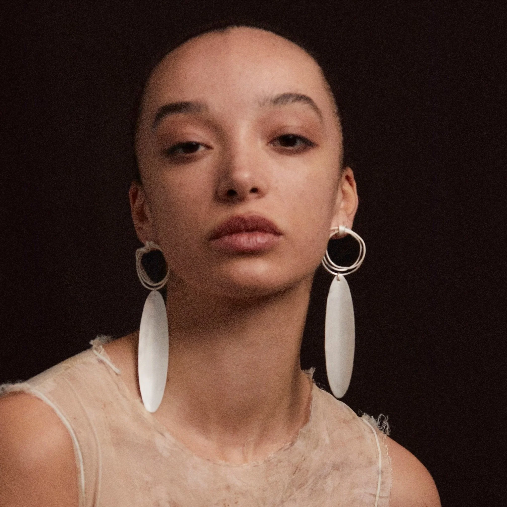 Woman wearing large oval earrings and a textured shirt, against a dark background.
