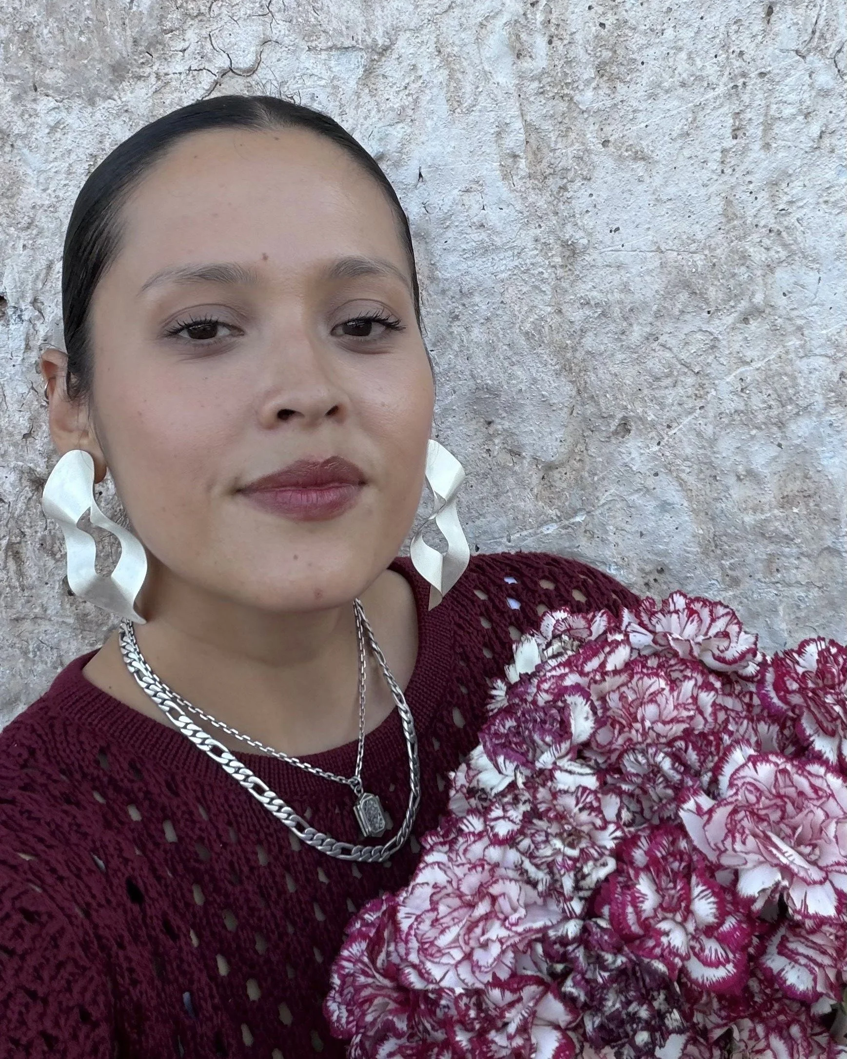 A woman with dark hair pulled back, wearing large white earrings, a silver chain necklace with a pendant, and a red crochet top. She is holding a bouquet of pink and white flowers, standing in front of a textured beige wall.