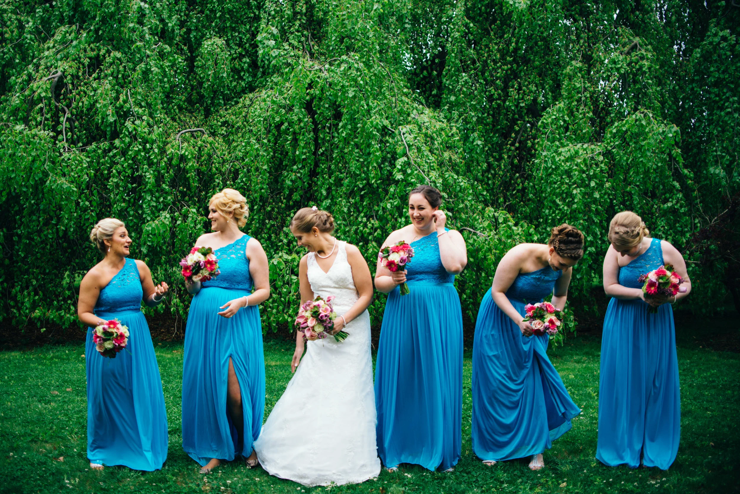 candid wedding photo of bridesmaids wearing blue one-shoulder dresses with pink bouquets