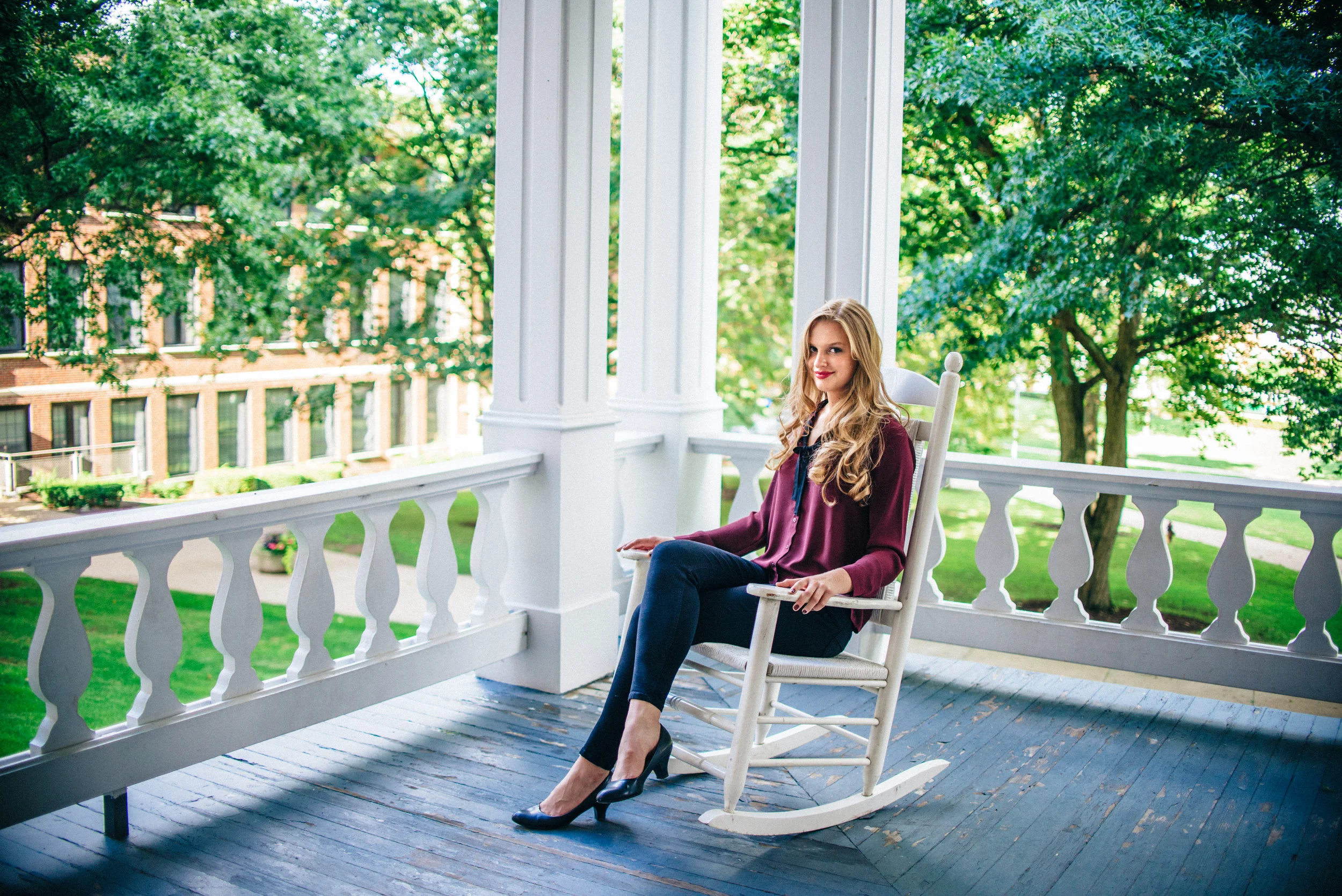 Blonde senior model in maroon blouse sitting on white rocking chair on the porch of Sutton Hall at Indiana University of Pennsylvania.