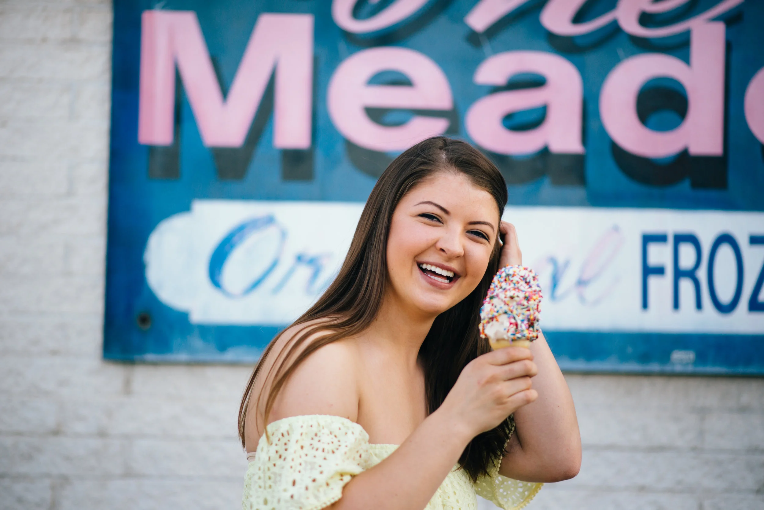 high school senior model with yellow lace off the shoulder top holding melting ice cream with rainbow sprinkles in front of the meadows ice cream store in Indiana, PA