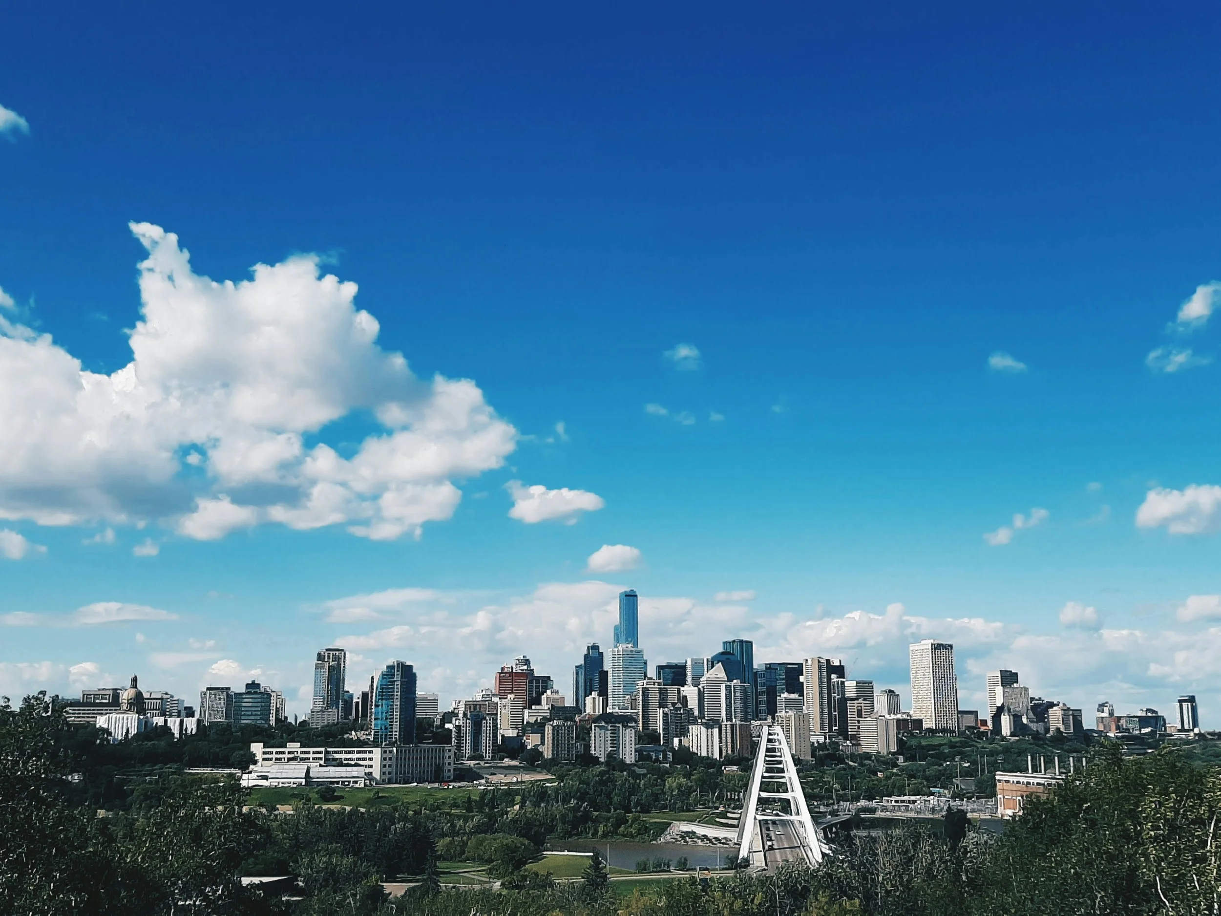 A city skyline with tall buildings under a blue sky with clouds. A white bridge runs through the center of the image.