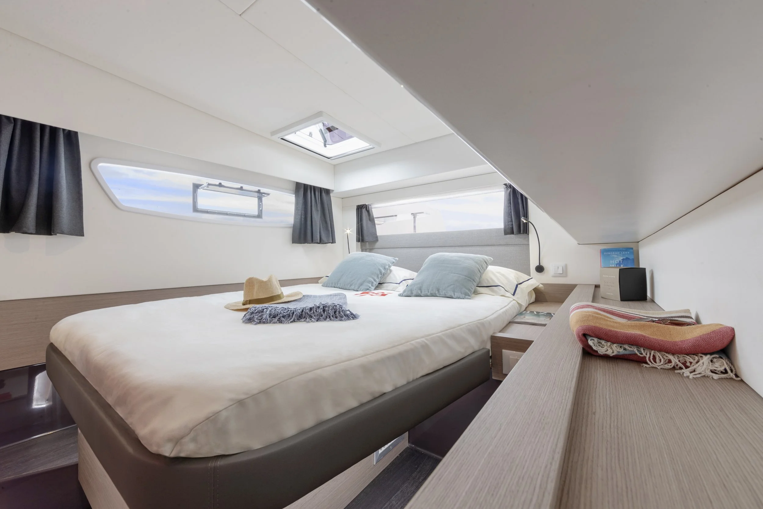 Interior of a modern boat cabin with a large bed, pillows, a hat, and a blanket, surrounded by windows with curtains, and a skylight overhead.