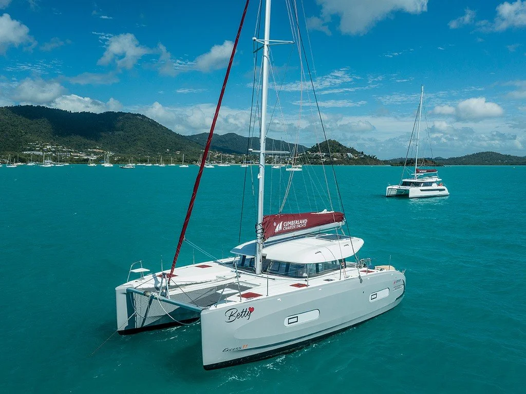 A white catamaran sailboat named Betty with a red sail cover on calm turquoise water, with another sailboat in the background, mountains, and a partly cloudy sky.