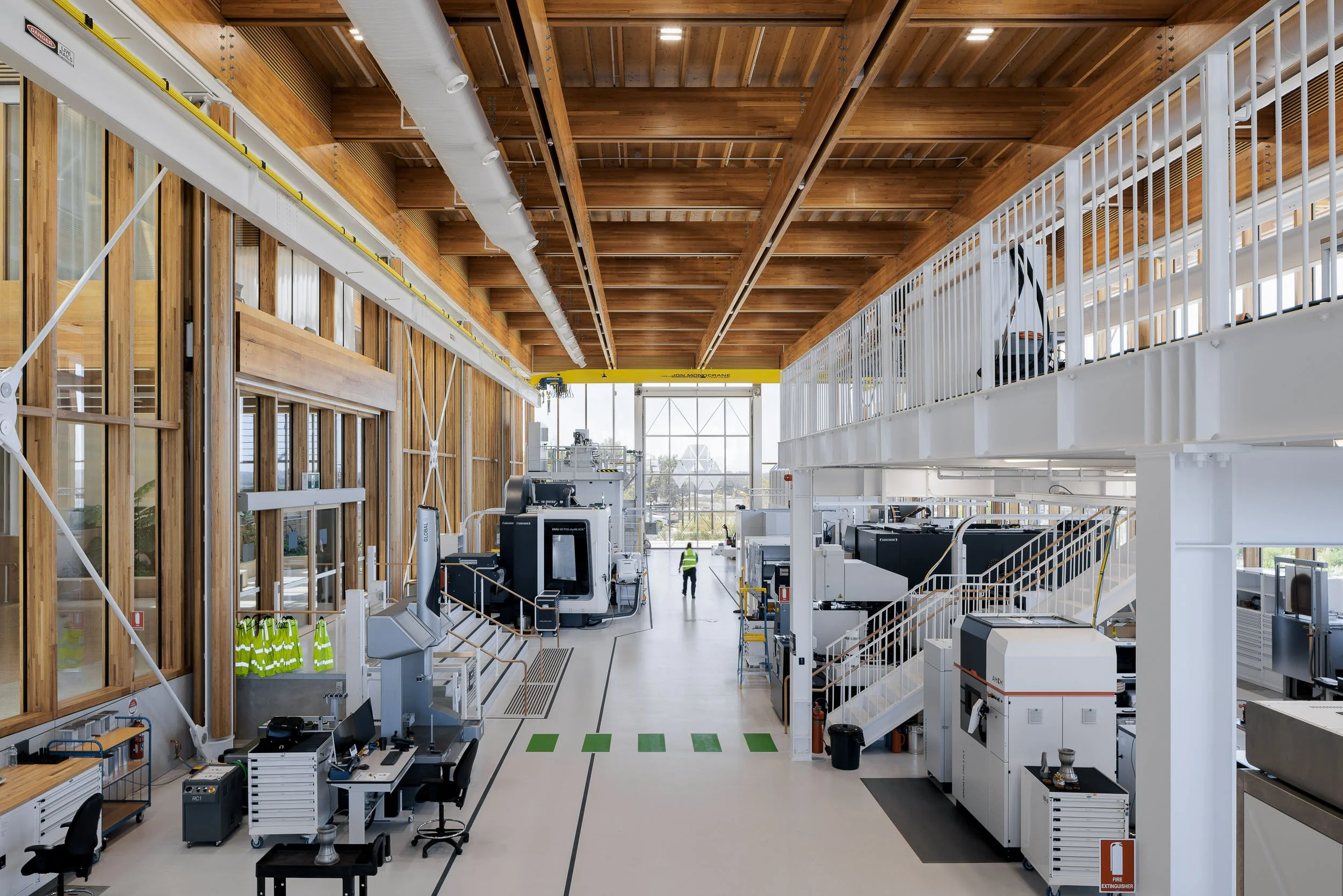 Interior of a modern industrial or laboratory facility with various equipment, machinery, and workstations, including a person in a safety vest near the large glass window at the back.