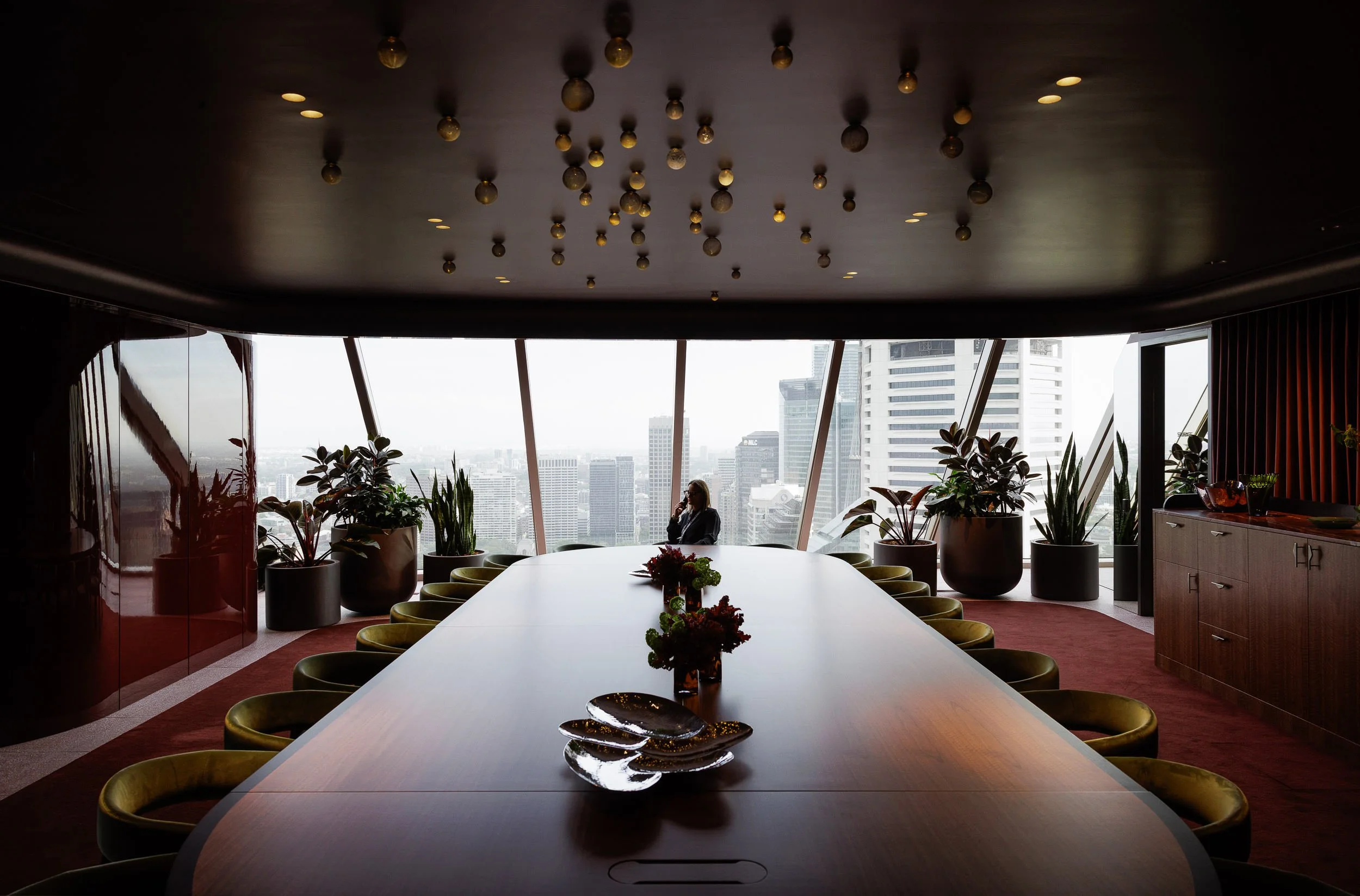 Cityscape view from a modern conference room with large windows, potted plants, a long conference table, and a woman sitting by the window on the phone.