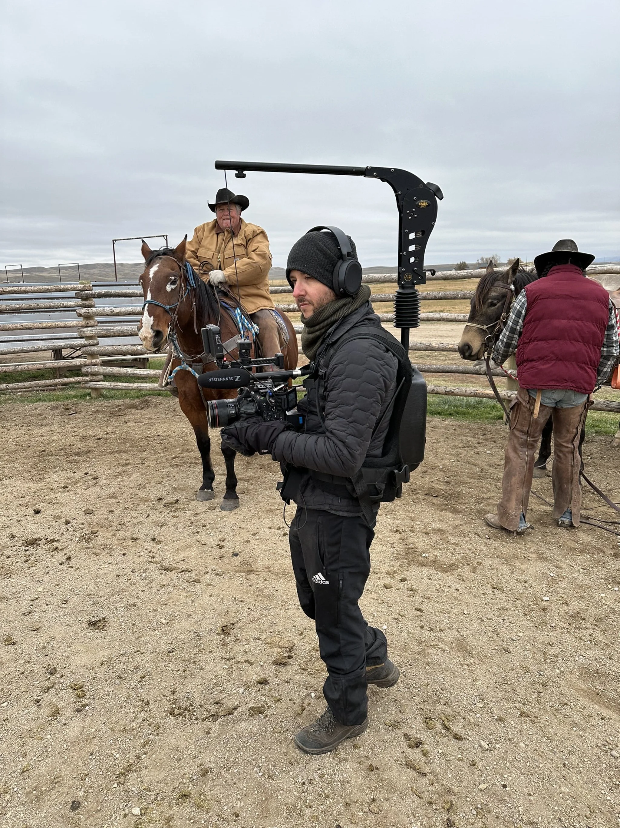 A cameraman films a man horseback riding inside a fenced outdoor area, with another person holding a horse nearby. The background shows a cloudy sky and open landscape.