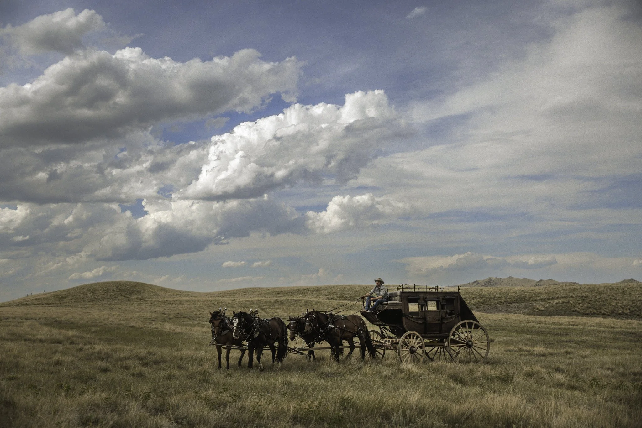 A stagecoach travels across a wide open prairie, pulled by four horses under a vast sky. Soft clouds stretch above the landscape, shaping the light and atmosphere. The scene reflects a slower, enduring way of life connected to land, movement, and tim