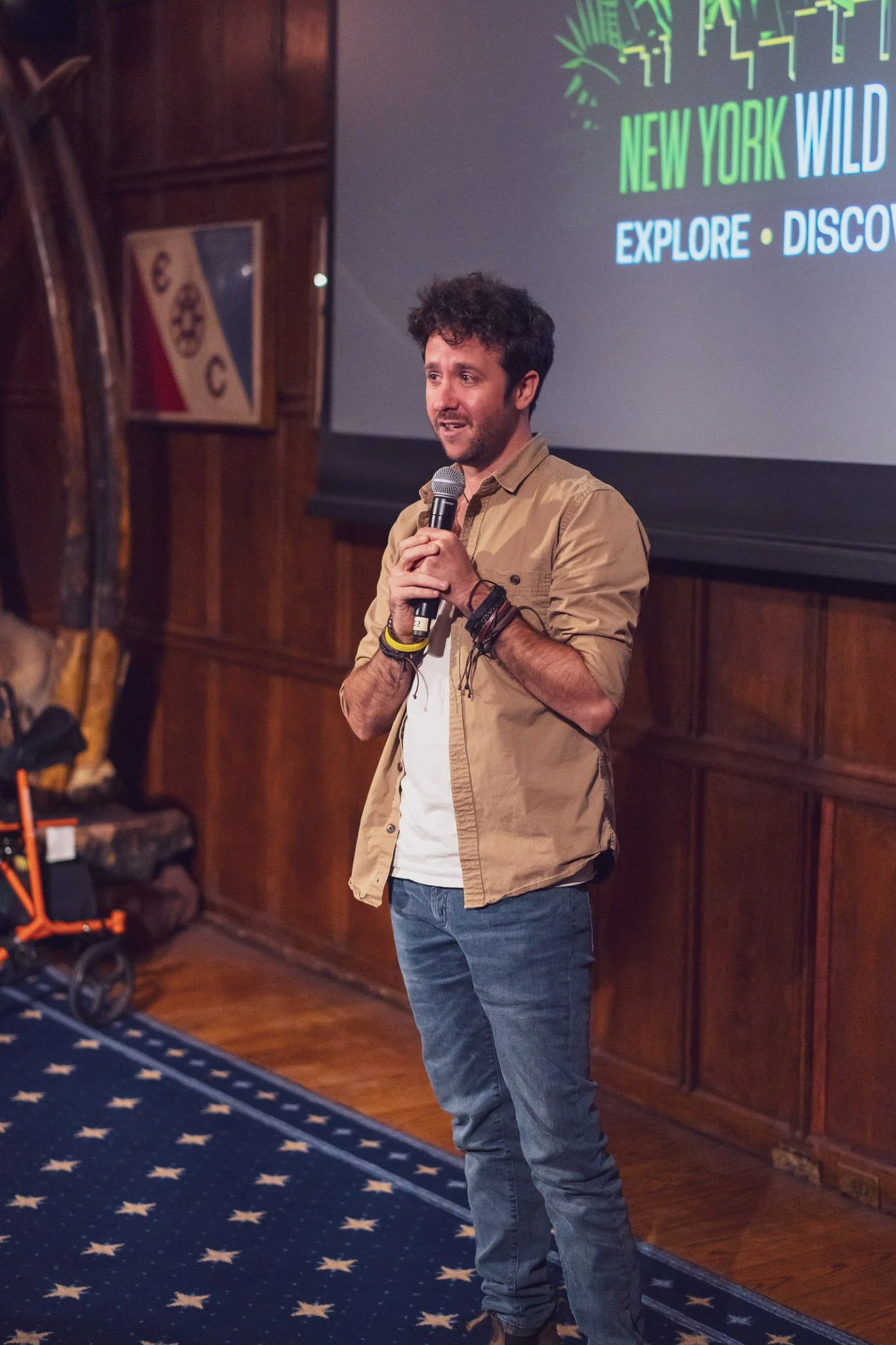 A man speaking into a microphone in front of a screen that reads "New York Wild Explore Discover" at an indoor event.