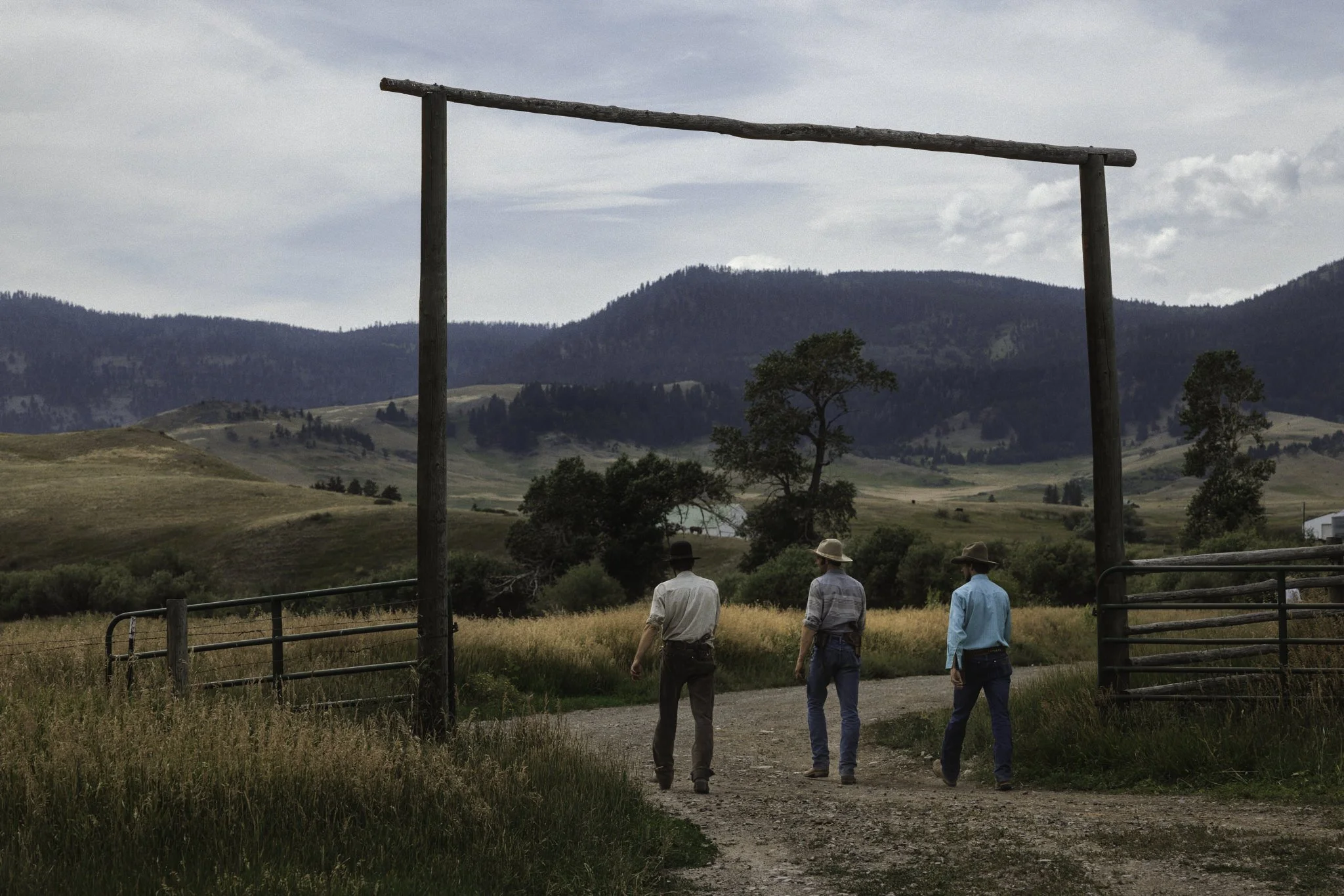 Three men walk through a ranch gate into a wide valley surrounded by rolling hills. The landscape reflects rural life shaped by land, routine, and a deep connection to place.