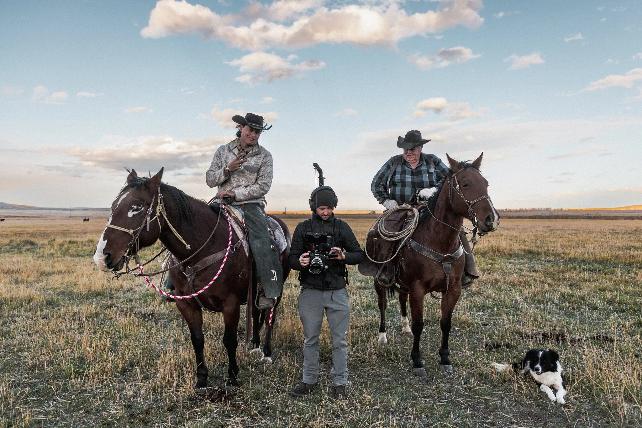 Two men on horseback in a grassy open field, with a camera operator standing between them holding a camera, and a black and white herding dog resting on the ground nearby under a partly cloudy sky.