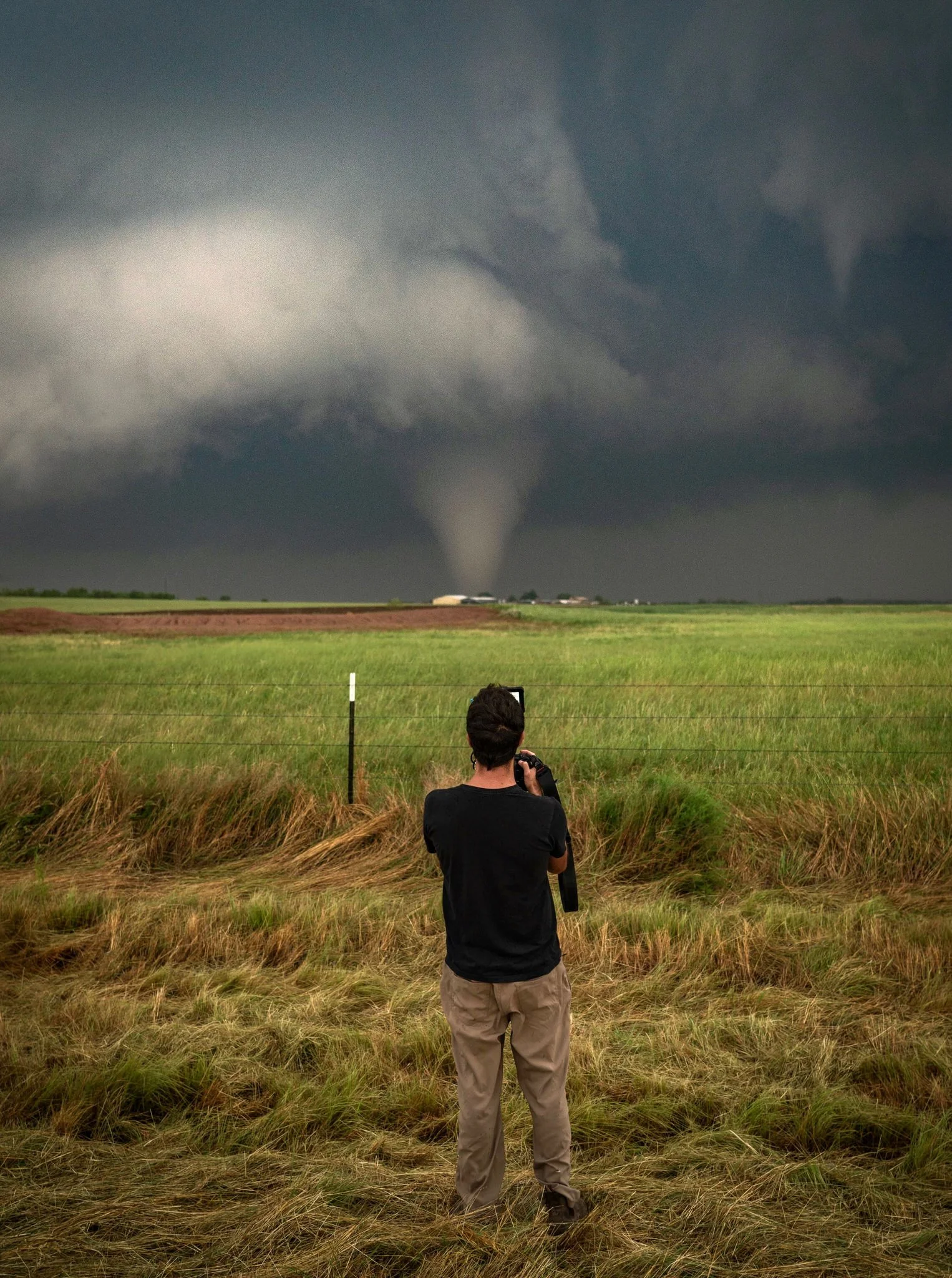 A person standing on a grassy field taking a photograph of a tornado in the distance under dark, stormy clouds.